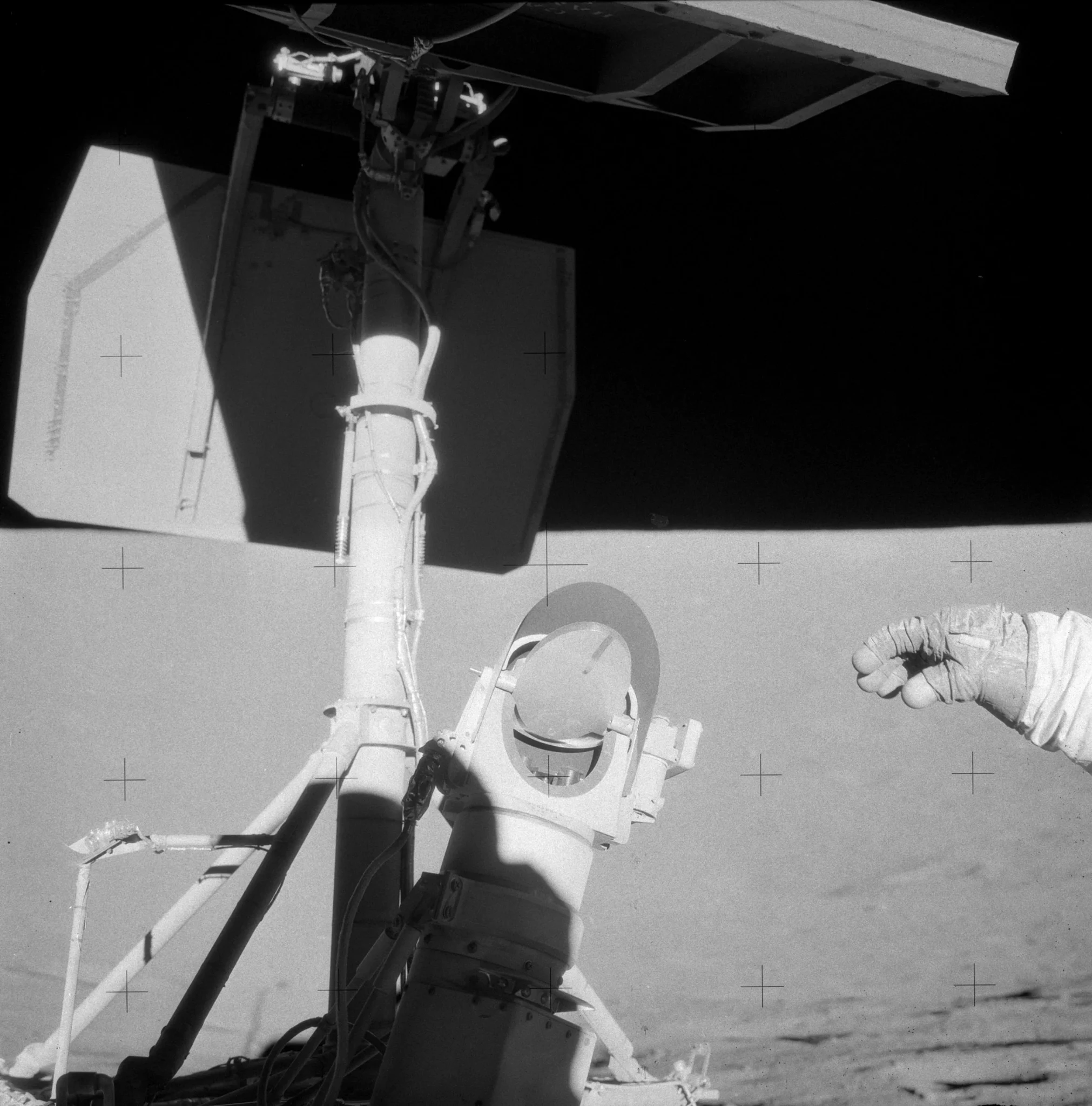 A large vertical pole holds metal panels aloft to the left of frame. In the center, a scientific instrument with a mirror covered in dust has had a single finger drawn across it, showing just how dusty it is. To the right of frame is an astronaut’s <br />outstretched hand. https://www.nasa.gov/history/alsj/picture.html