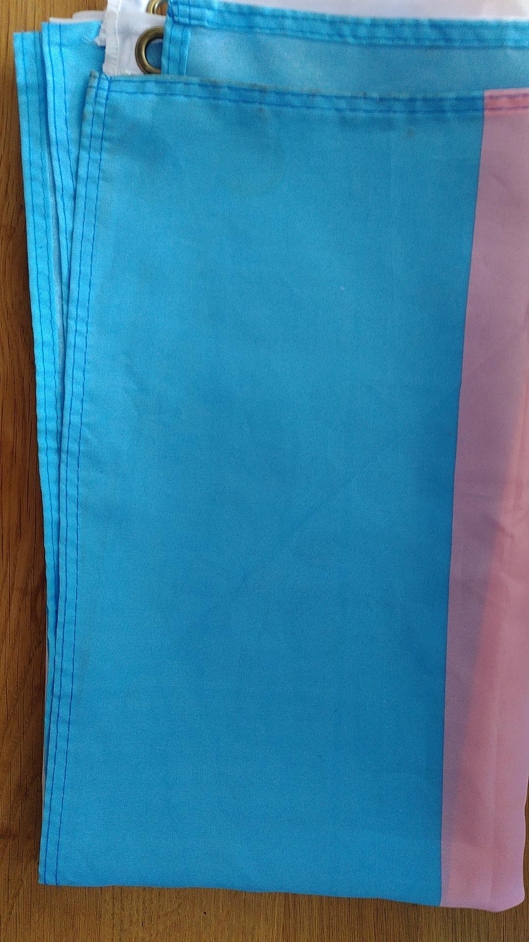 Zusammengefaltete Trans-Flagge
Siehtbar ist links ein blauer und rechts daneben ein rosaner Abschnitt. Im Hintergrund ist noch eine Öse zu sehen.